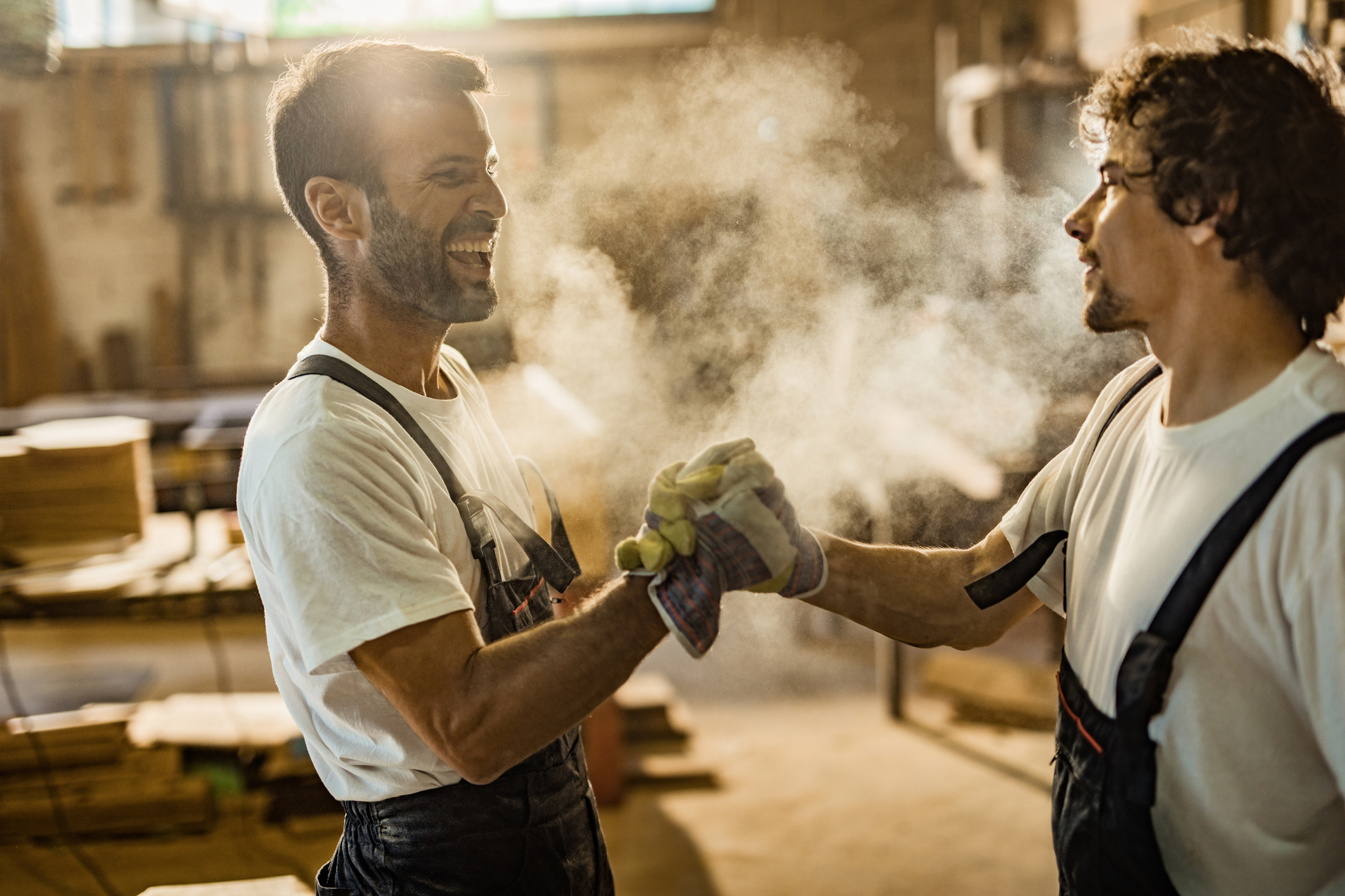 Happy male carpenters greeting each other while holding hands in a workshop.